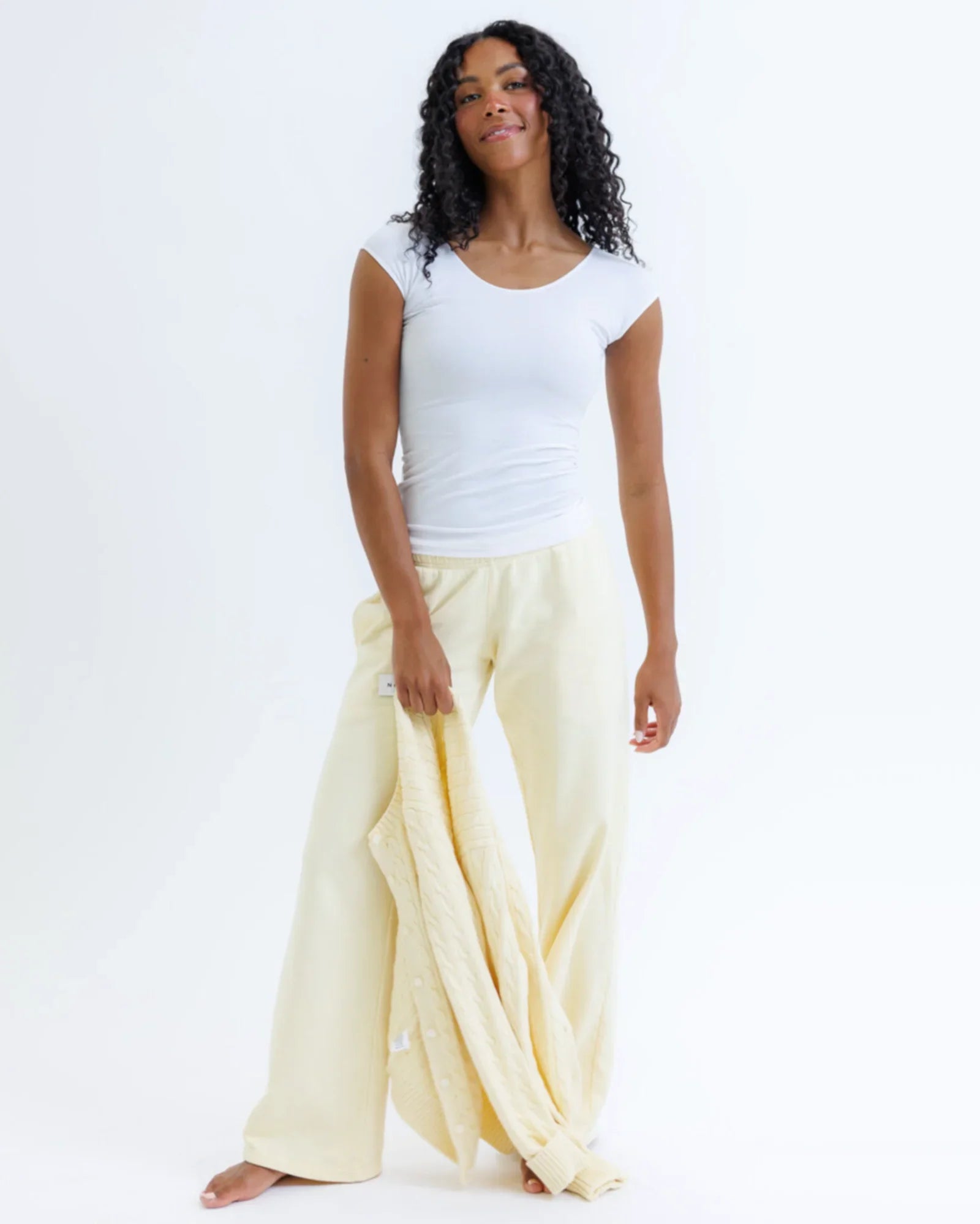 Woman wearing fitted white cap sleeve t-shirt and cream lounge pants, holding sweater